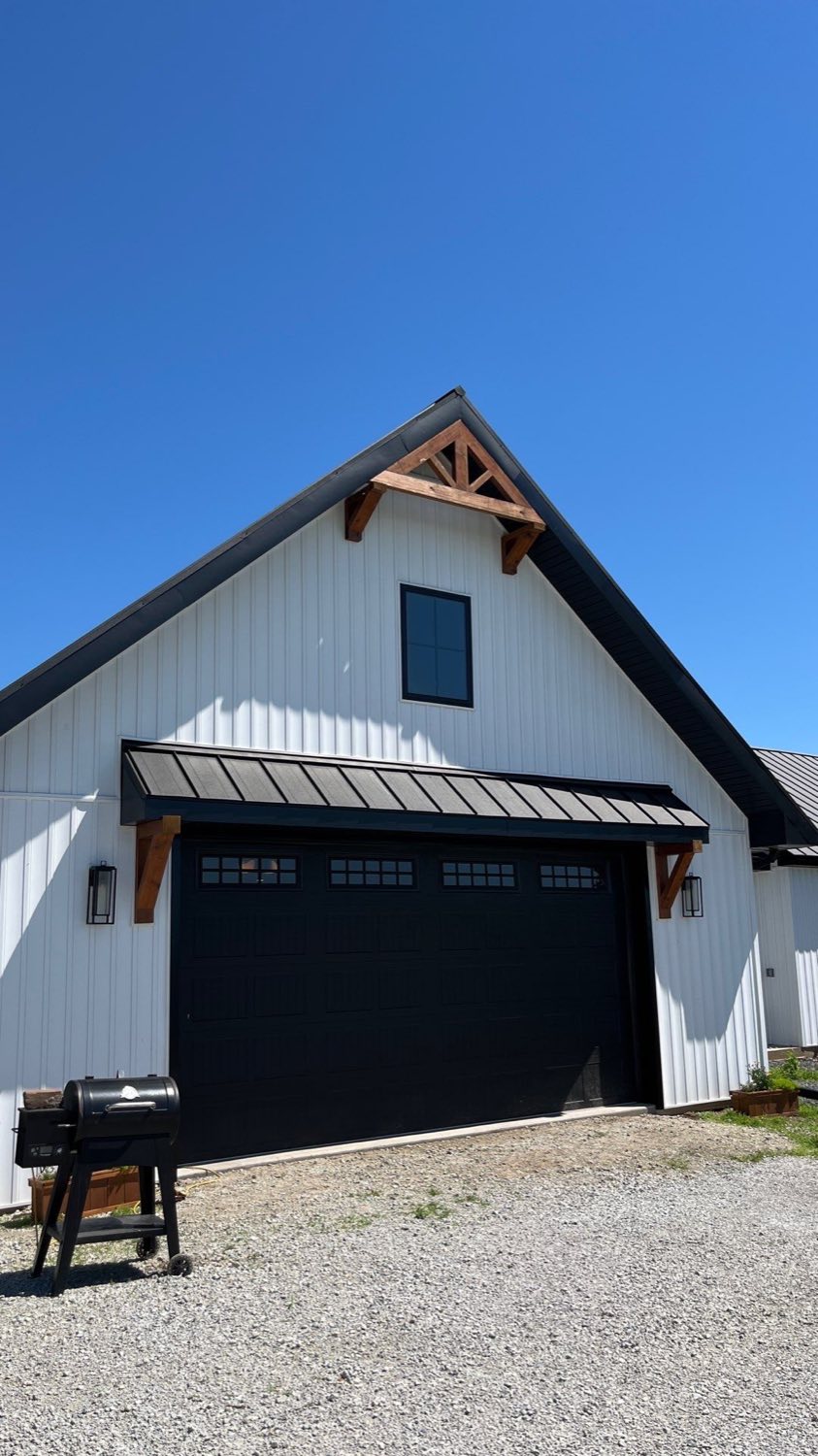 White board-and-batten farmhouse with a black standing-seam roof, timber truss in the gable, and a black carriage-style garage door