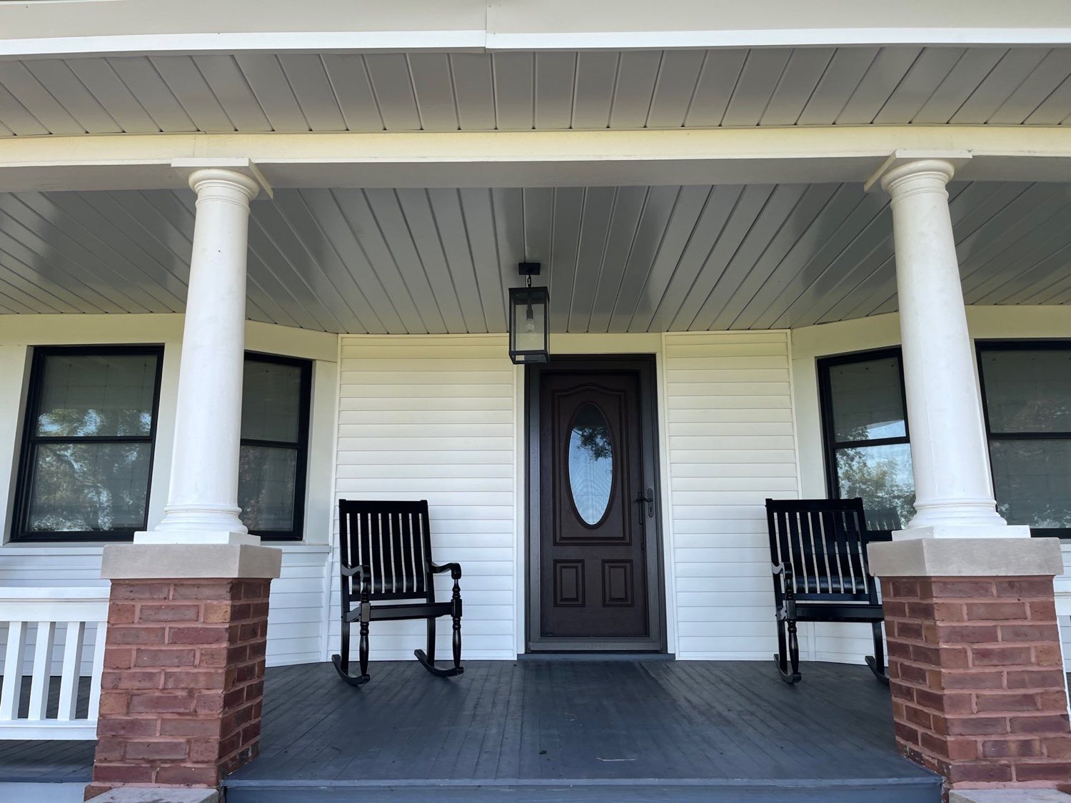 Restored farmhouse porch with white columns, a beadboard ceiling, brick piers, and wooden rocking chairs by the front door